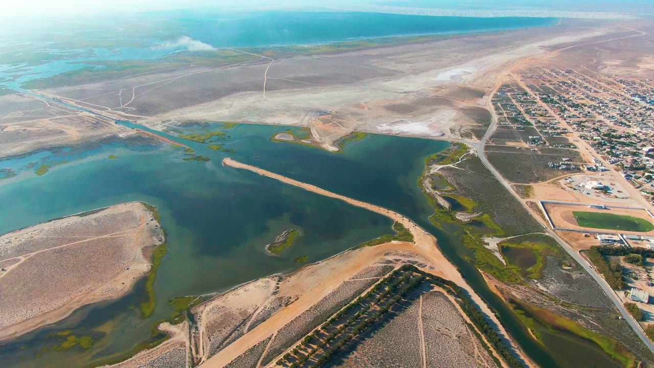 High aerial view of calm water crossed by a narrow bar beside dry terrain and town streets.