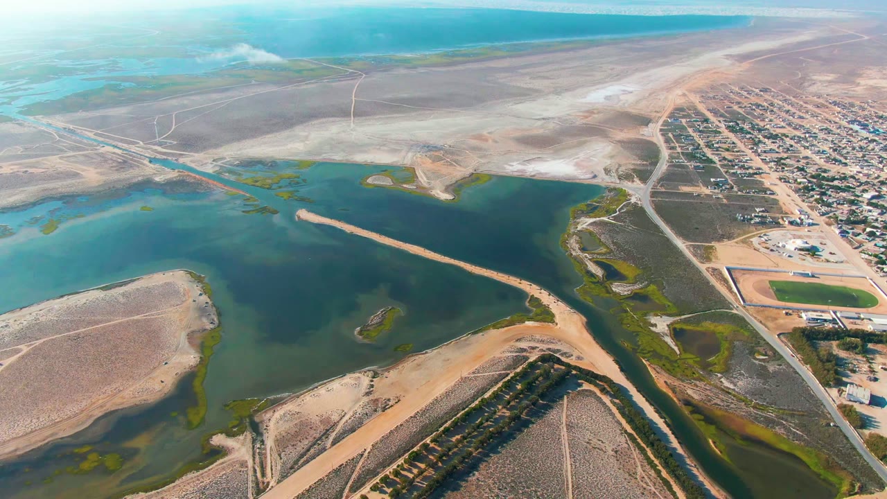 Guerrero Negro aerial frame with the water basin filling the left side, tan arid ground in front, a road network and town edge to the right, and a pale smoke or steam plume near the horizon, no visible people.
