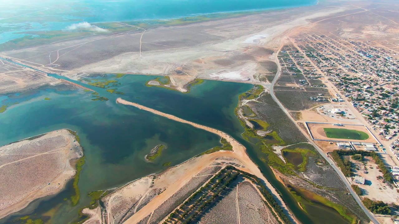 A high aerial view over Guerrero Negro shows blue-green lagoon water, pale sandbars, brown flats, roads, and a small settlement to the right, with no visible people.