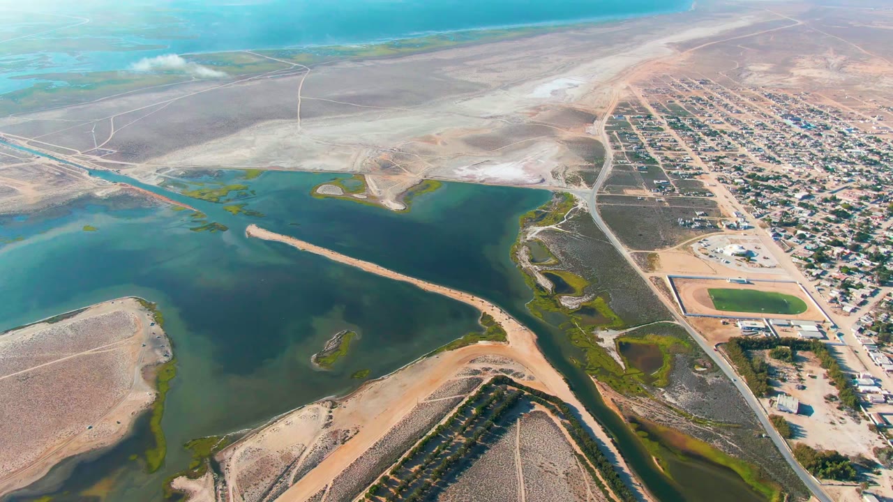 An aerial frame over Guerrero Negro shows darker lagoon channels around pale sand, green shoreline patches, and a dense grid of buildings on dry land, with no visible people.