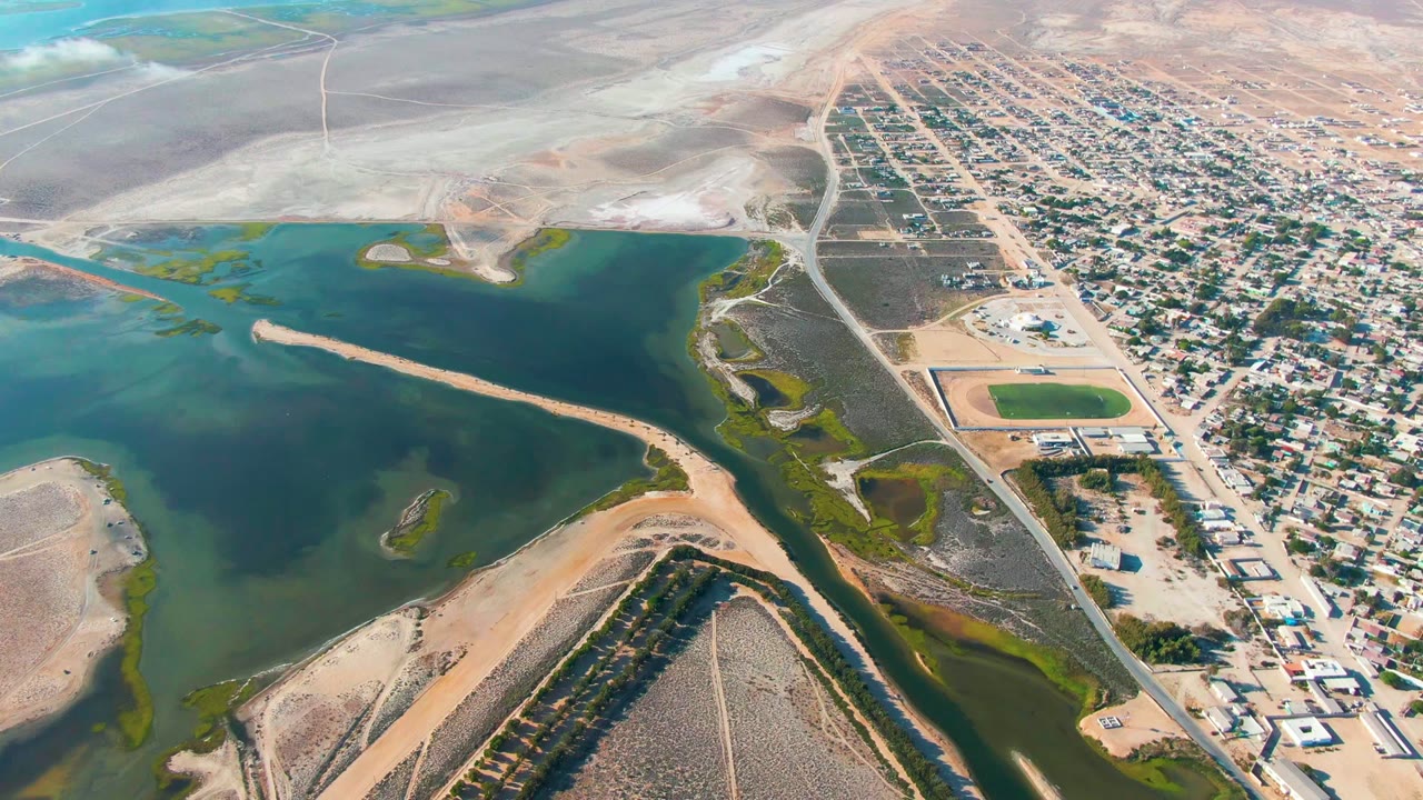 Drone view of a lake edge, sandy causeway, houses, and roads.