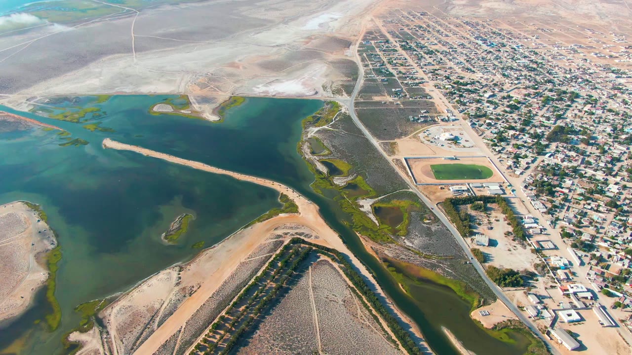 Aerial frame in Guerrero Negro showing more of the residential grid beside blue-green water, pale dry land, shoreline vegetation, and no visible people.