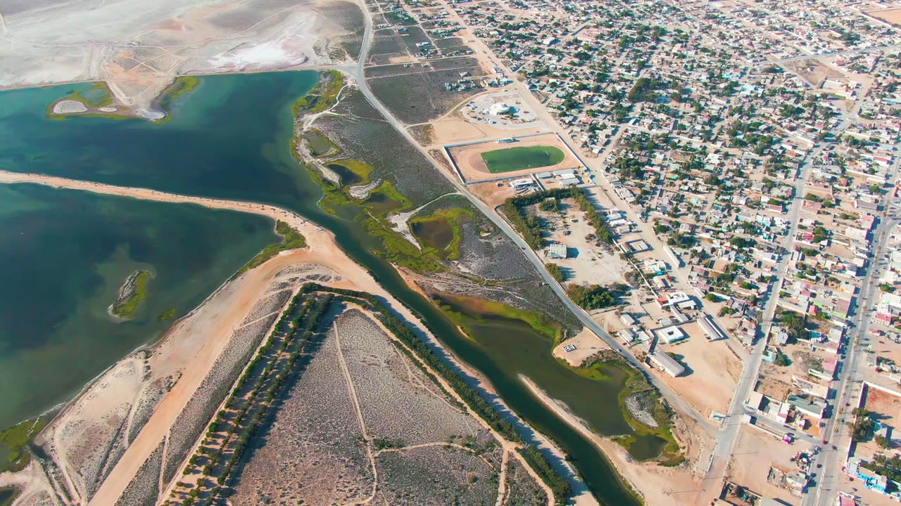High aerial view over Guerrero Negro, Baja California Sur, showing blue-green lagoon water, a sandy spit, roadways, sports fields, and dense low residential blocks with no visible people.