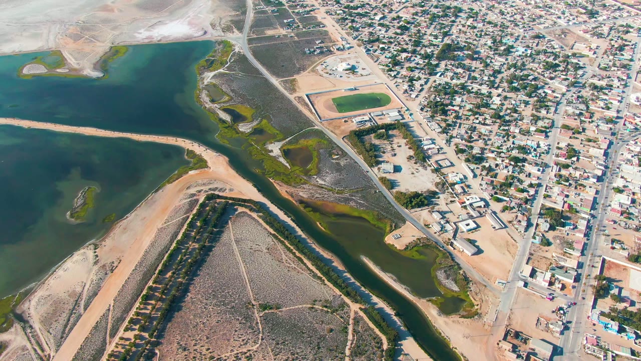 Aerial view over Guerrero Negro where blue water and light sand occupy the left and lower frame while compact houses, straight roads, and a pair of rectangular sports fields sit to the right, with no visible people.