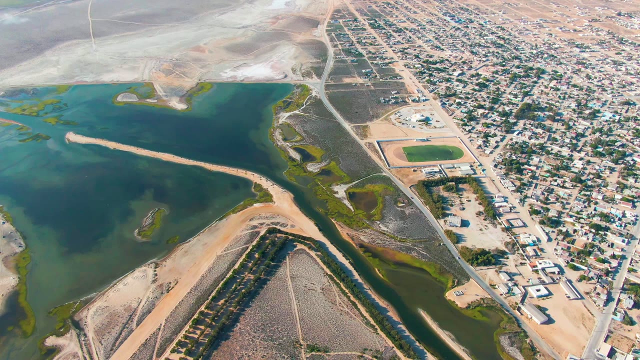 Aerial frame over Guerrero Negro, Baja California Sur, showing green-blue water on the left, sandy banks and a canal below, residential blocks to the right, and no visible people.