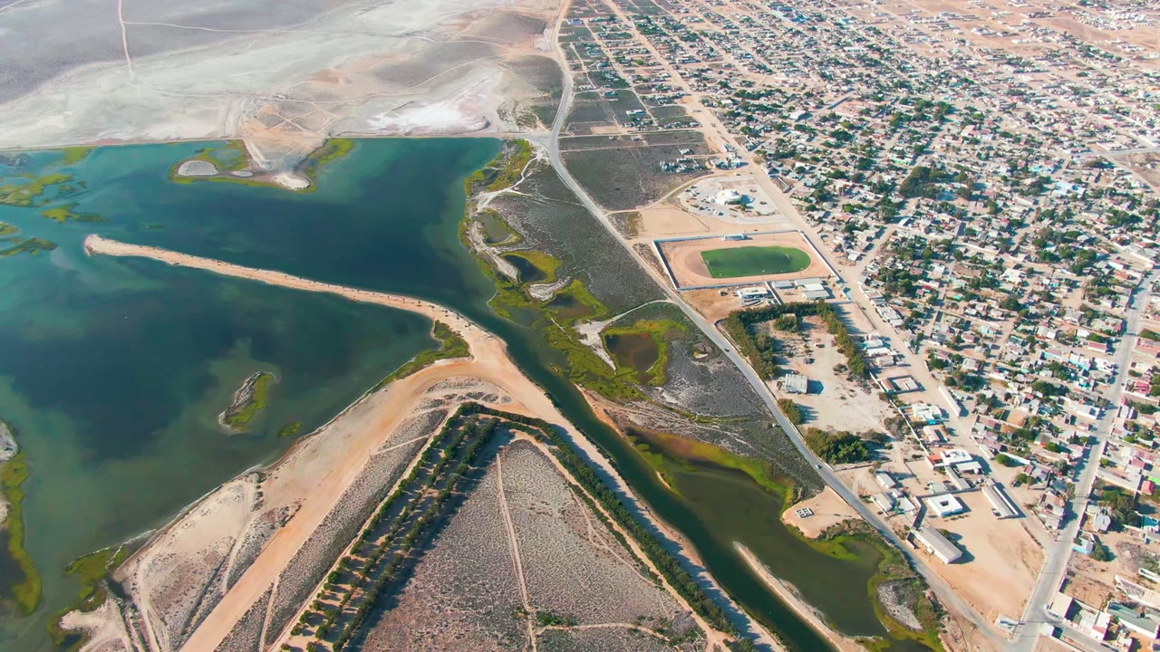 Wide aerial frame over Guerrero Negro showing green water and sandy banks at left, a dark channel below, pale desert ground at top, a residential street grid at right, and no visible people.