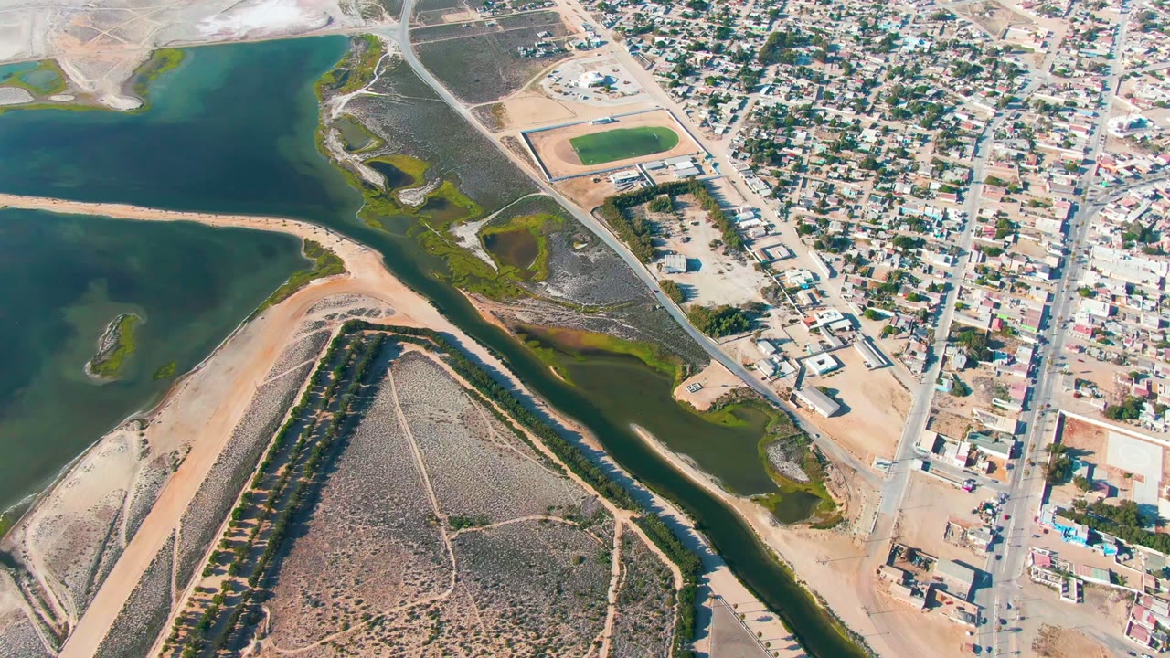 High aerial view over Guerrero Negro, Baja California Sur, with green-blue water channels, a sandy spit, dry scrubland, town blocks, roads, and no visible people.