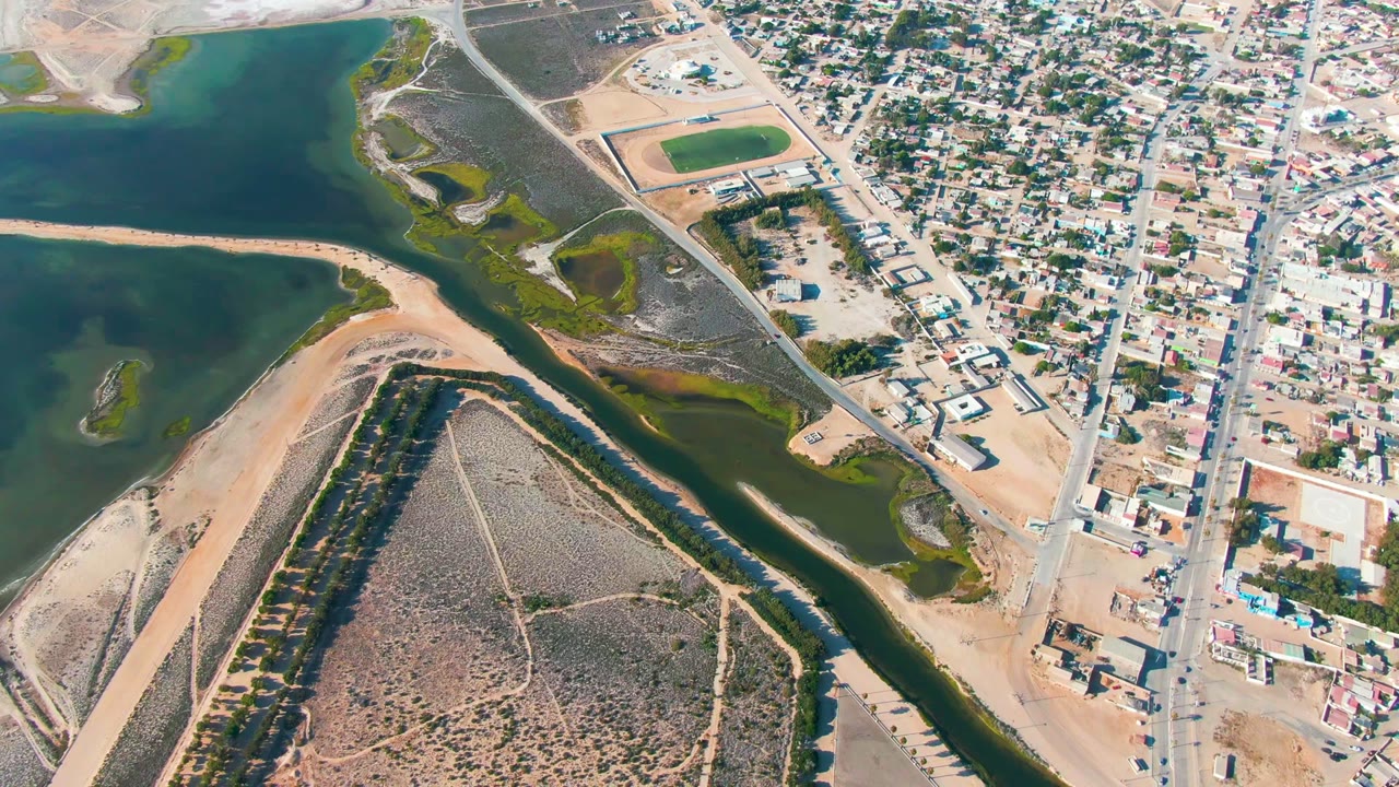 Drone view over Guerrero Negro with a broad green water area at left, a narrow channel through the center, sandy open land below, dense streets at right, and no visible people.