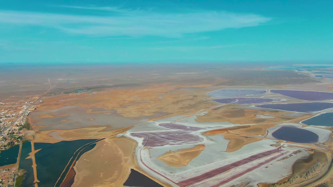 Wide aerial view over Guerrero Negro salt evaporation ponds, with purple and blue basins, pale salt flats, brown dry land, a road and settlement at left, and no visible people.