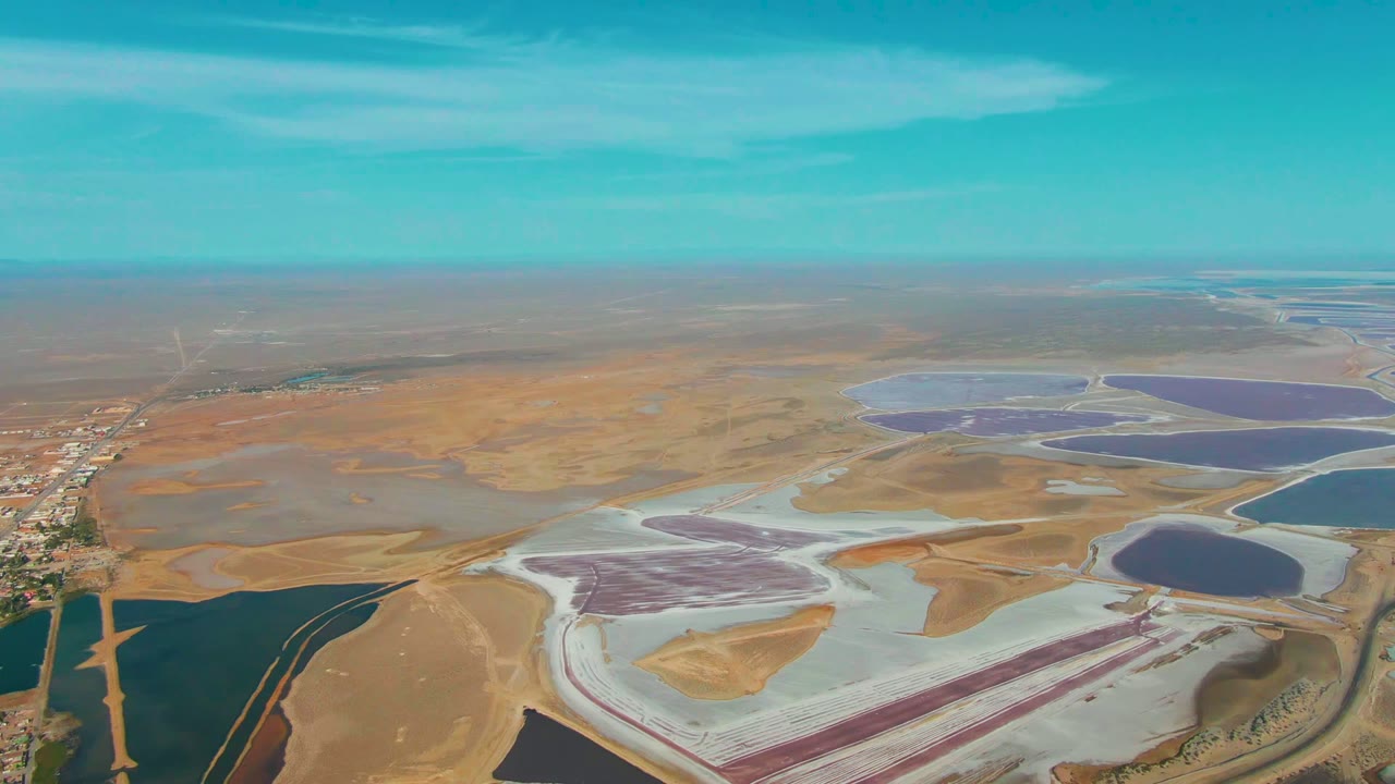 Guerrero Negro aerial view with a large pale salt area and purple basin near the lower center, blue evaporation ponds to the right, dry brown earth across the background, and no visible people.
