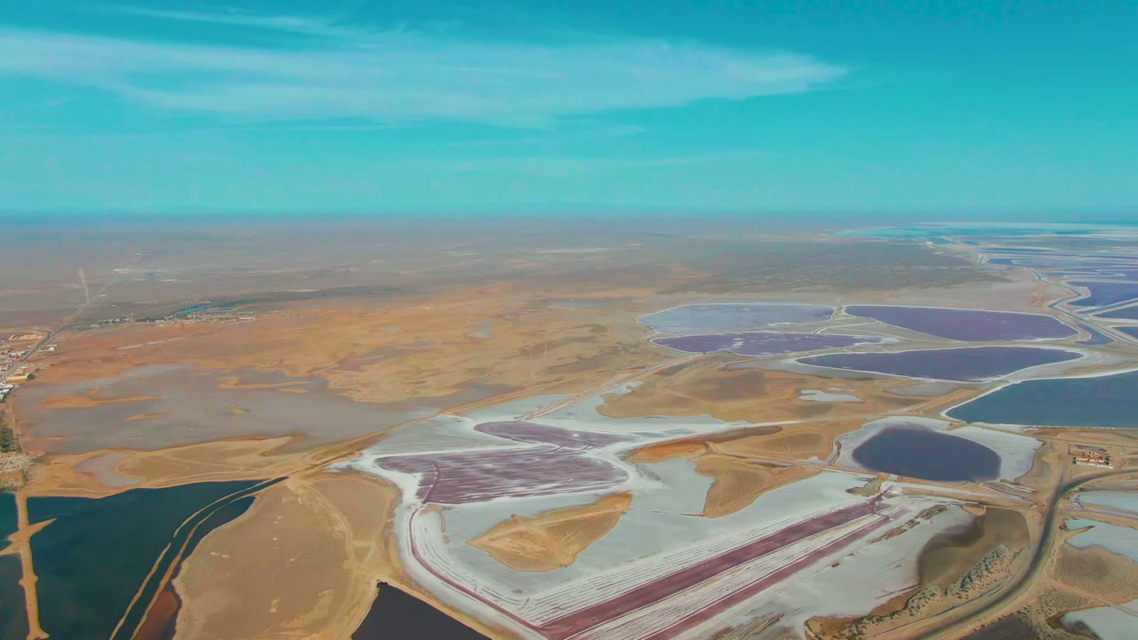 Wide aerial frame over Guerrero Negro salt evaporation ponds, with pale salt flats, purple and blue water basins, tan desert, turquoise sky, and no visible people.