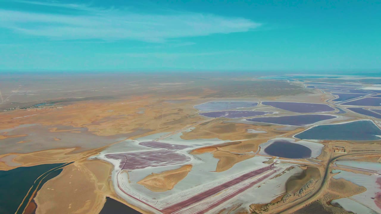 Opening aerial frame over Guerrero Negro, Baja California Sur, showing pink, purple, blue, and pale evaporation ponds across dry beige terrain with no visible people.