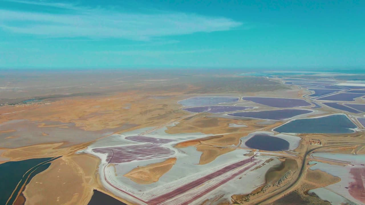 Oblique aerial view of Guerrero Negro salt evaporation ponds, with pale flats and reddish-purple water in the foreground, blue basins to the right, distant pond grids, and no visible people.