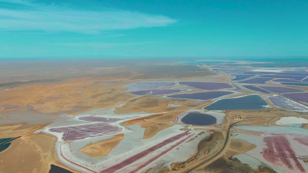 Guerrero Negro aerial still with geometric evaporation ponds, white salt margins, a dark blue basin near center, tan desert beyond, and no visible people.
