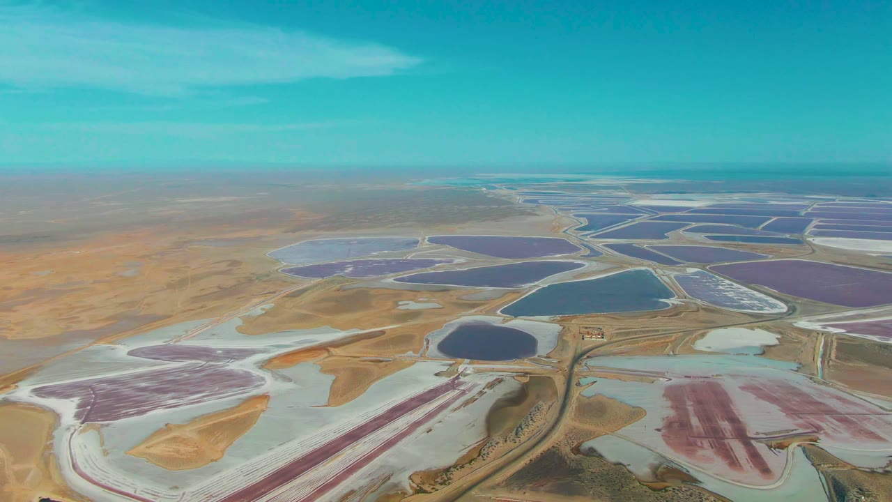Aerial view over Guerrero Negro salt evaporation ponds with pink and white flats in the foreground, darker blue ponds beyond, a flat horizon, and no visible people.