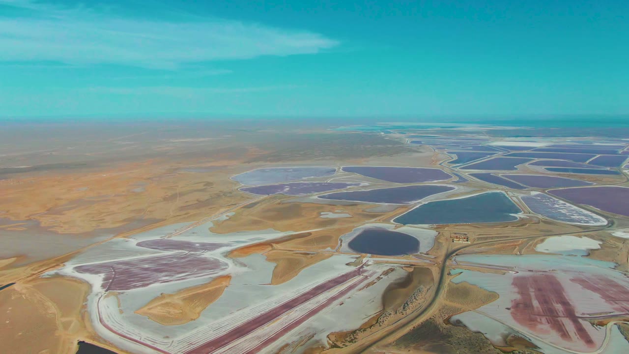 High aerial view over Guerrero Negro salt evaporation ponds, with pink and purple basins, white salt flats, blue water, dry tan land, a low horizon, and no visible people.