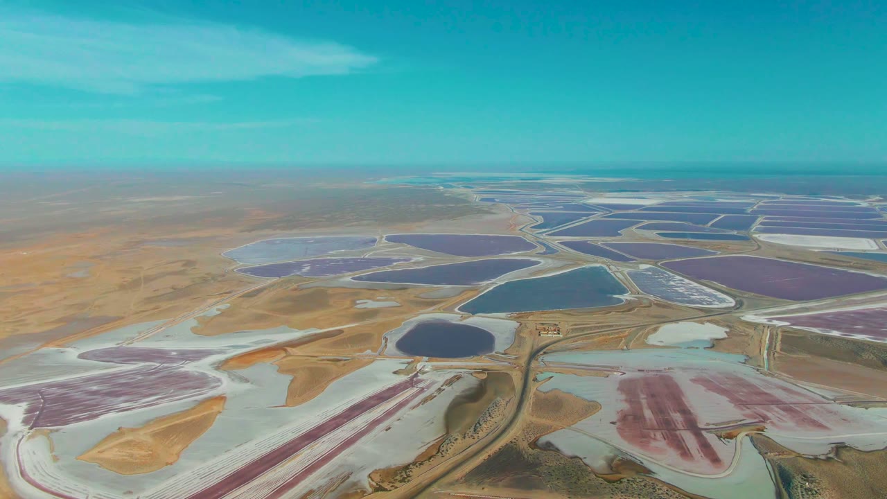 Opening aerial frame over Guerrero Negro salt evaporation ponds, with purple, pink, blue, and white pond sections spread across tan dry land and no visible people.