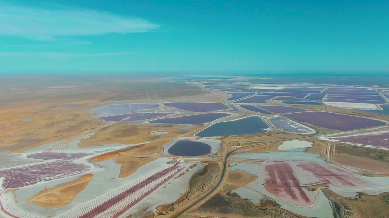 Aerial view across Guerrero Negro salt ponds with pale white flats in the foreground, darker blue ponds near center, purple ponds to the right, and no visible people.