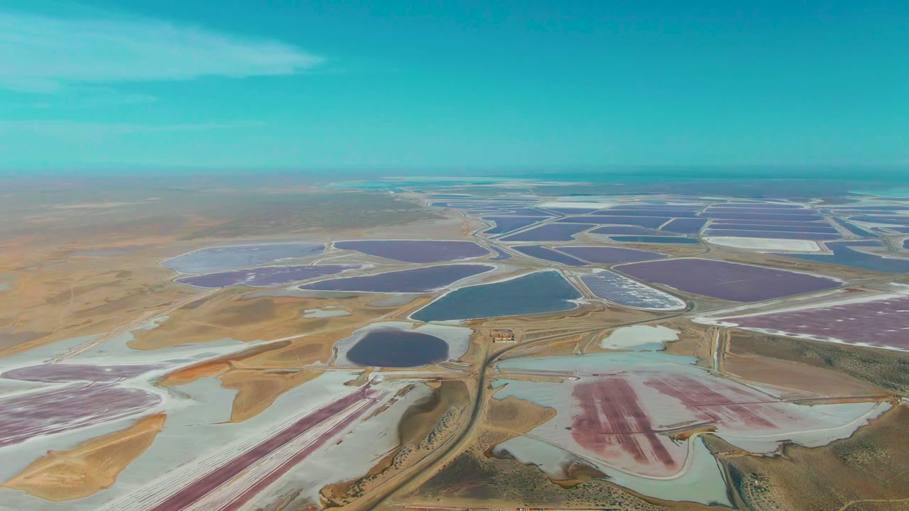 Aerial view over Guerrero Negro salt evaporation ponds with purple, pink, blue, and pale basins across tan arid terrain, with no visible people.