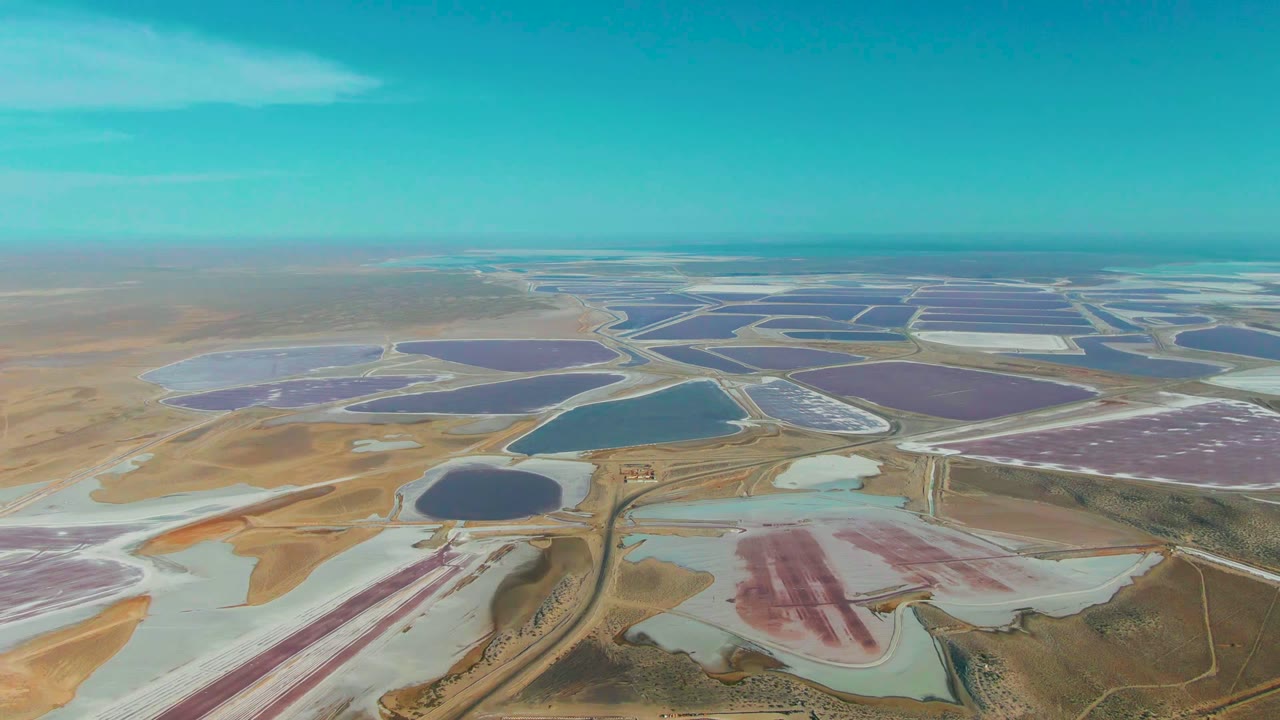 Opening aerial frame over Guerrero Negro salt evaporation ponds with pink, purple, blue, and pale basins spreading across dry terrain under a clear sky, no visible people.