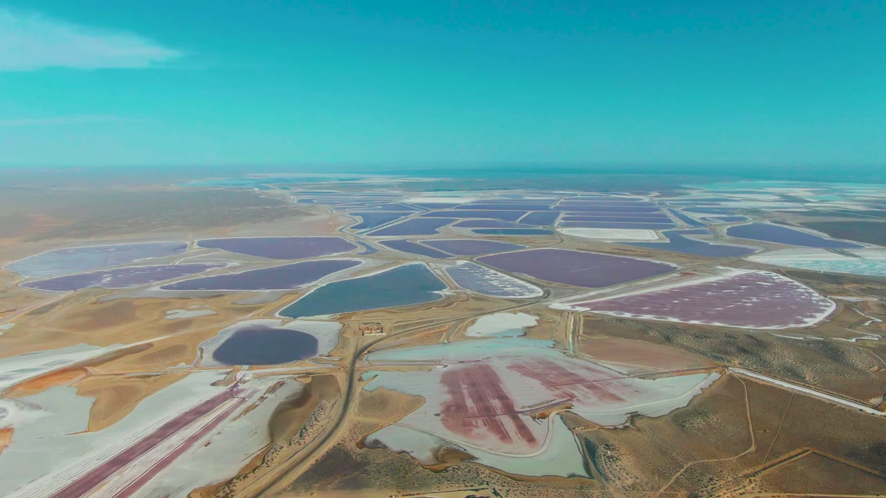 Aerial view over Guerrero Negro evaporation ponds, with pink, purple, blue, and pale basins separated by dirt roads and no visible people.