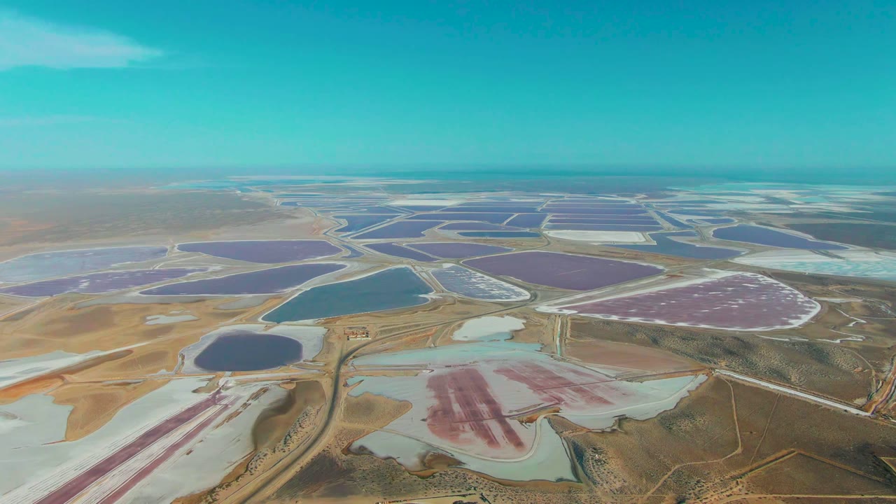 Wide aerial frame over Guerrero Negro salt ponds, with a blue basin near center, purple evaporation ponds in the distance, pale salt deposits, dirt tracks, and no visible people.