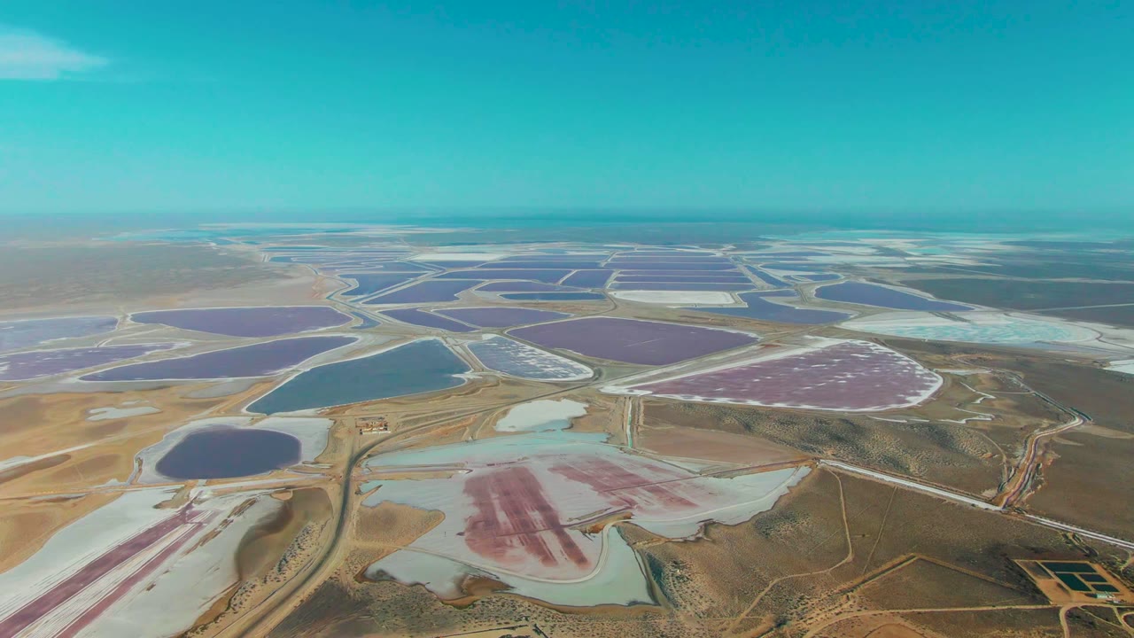 Aerial frame over Guerrero Negro with pale salt basins in the foreground, purple and blue evaporation ponds in the midground, service roads between them, and no visible people.