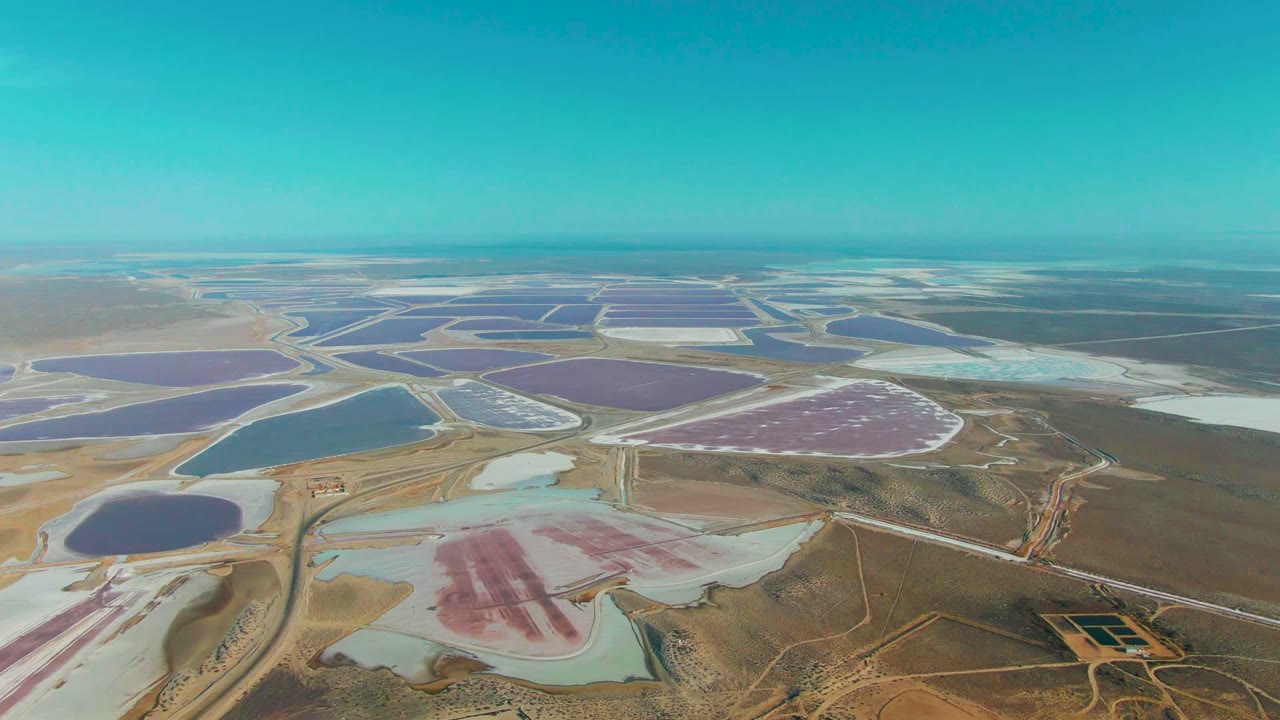 Wide aerial frame over Guerrero Negro salt evaporation ponds, with blue and pink basins separated by pale berms, dry terrain below, turquoise sky above, and no visible people.
