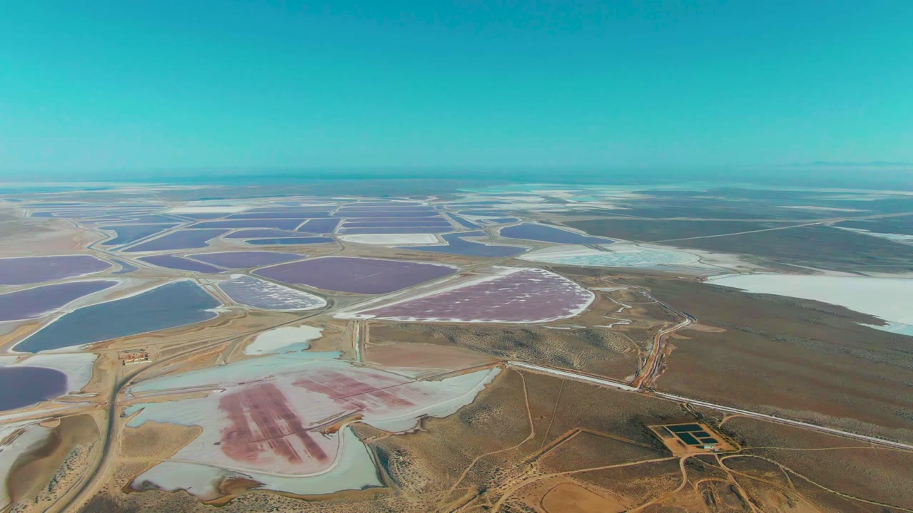 Drone view over Guerrero Negro evaporation basins where a pink salt pond sits near pale flats and larger purple-blue ponds extend toward the horizon, with no visible people.