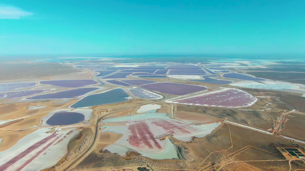 High aerial view over Guerrero Negro, Baja California Sur salt evaporation ponds, with pink and pale blue basins in the foreground, purple and blue ponds beyond, and no visible people.