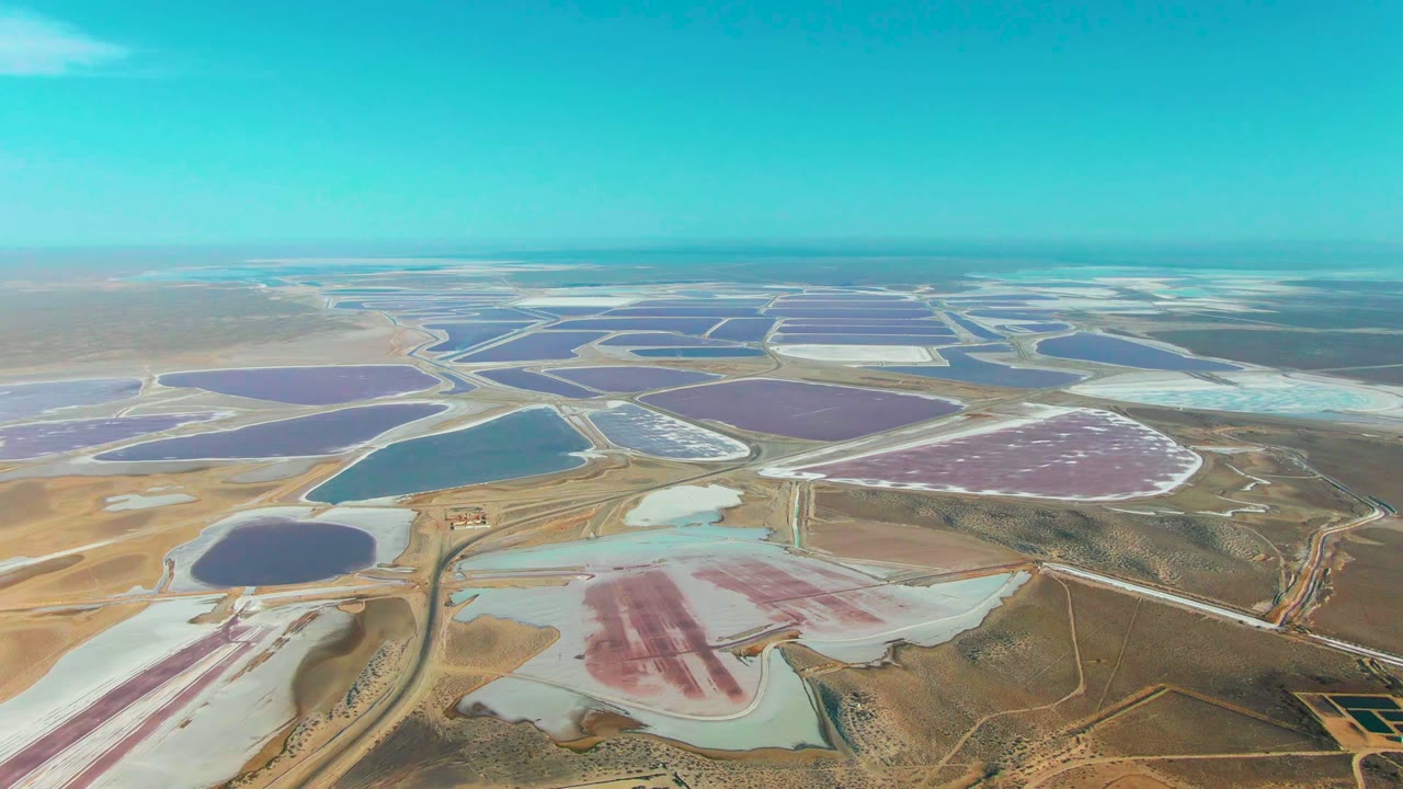 High view across Guerrero Negro salt ponds with angular water basins, pale salt flats, dusty roads, dry brown land, and no visible people.