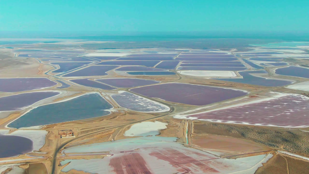 Aerial view over Guerrero Negro salt evaporation ponds, with purple, blue, white, and turquoise basins divided by tan roads and no visible people.