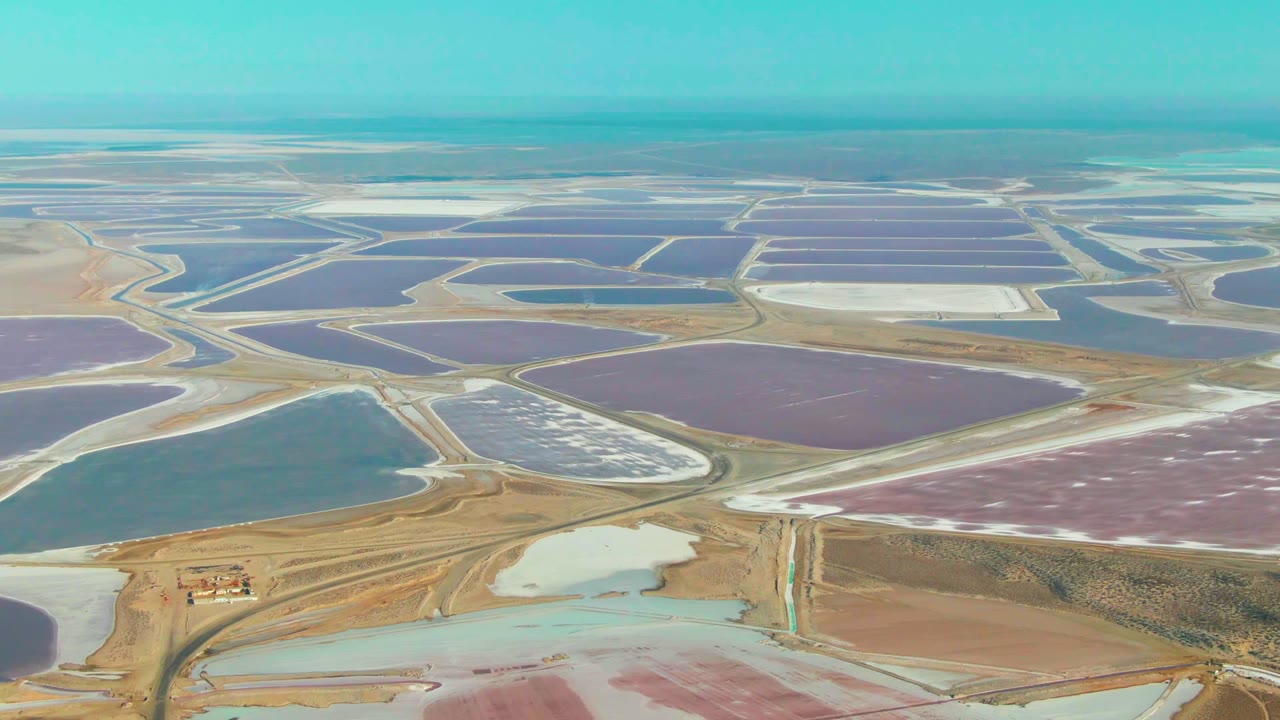 Aerial view across Guerrero Negro evaporation ponds, with a large purple basin, pale white pond sections, blue-green water, tan embankments, and no visible people.