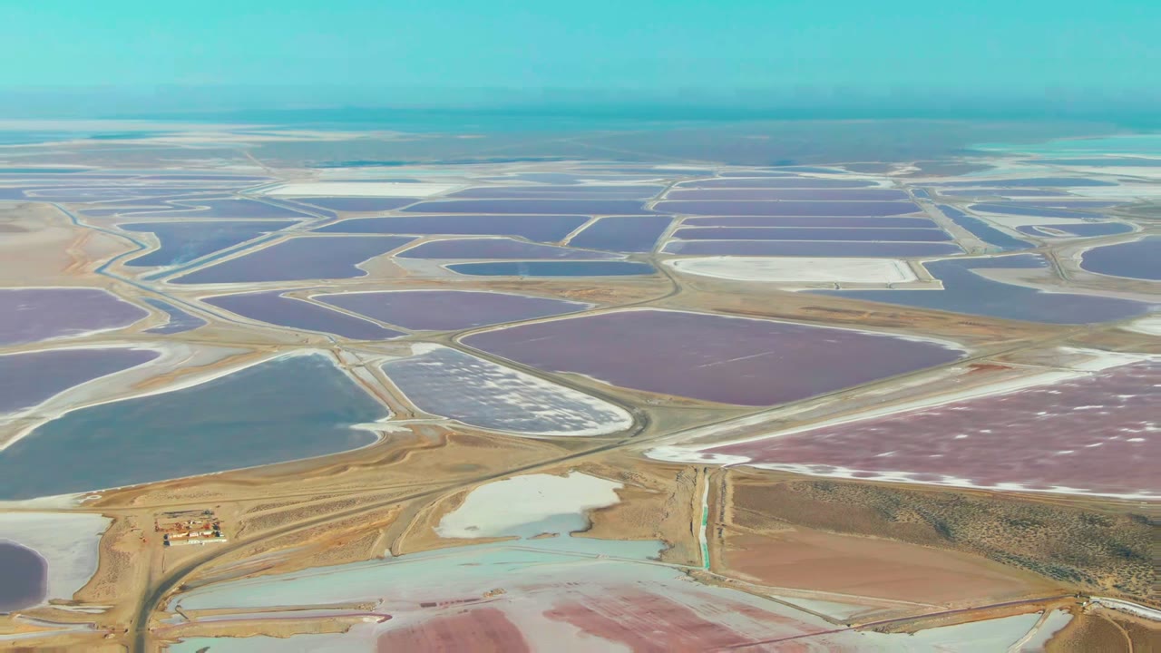 A high aerial view over Guerrero Negro salt evaporation ponds shows purple, blue, pink, and white basins divided by pale roads, with dry terrain and no visible people.