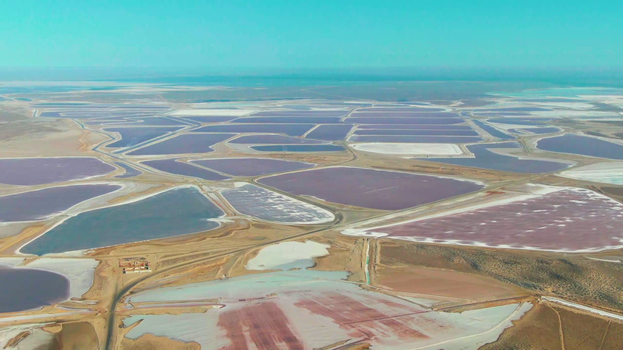 The Guerrero Negro aerial view holds on broad rectangular and irregular salt ponds, with blue and purple basins stretching toward a flat horizon and no visible people.