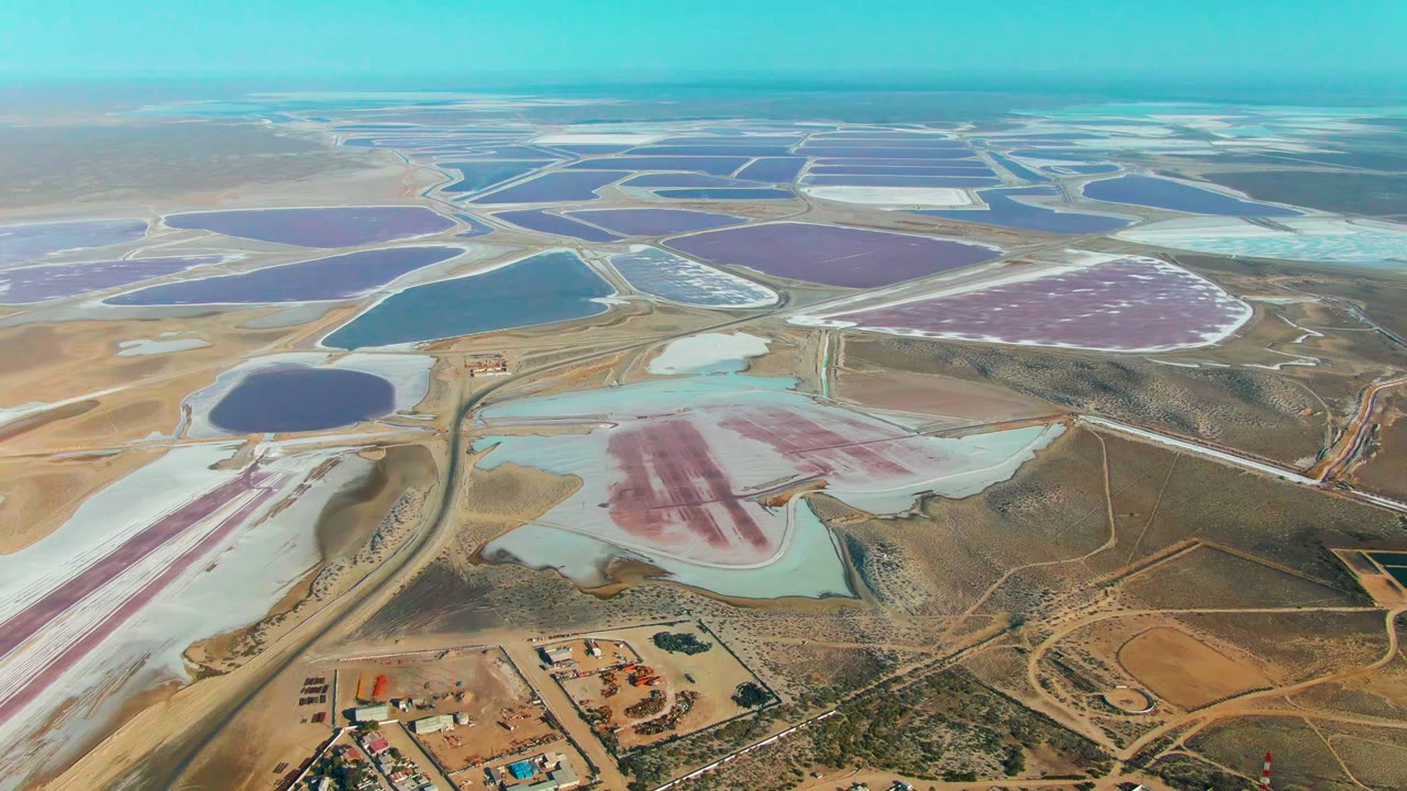 Aerial view over Guerrero Negro evaporation ponds, with angular purple and blue basins across the upper frame, white salt sections, dry roads, and no visible people.