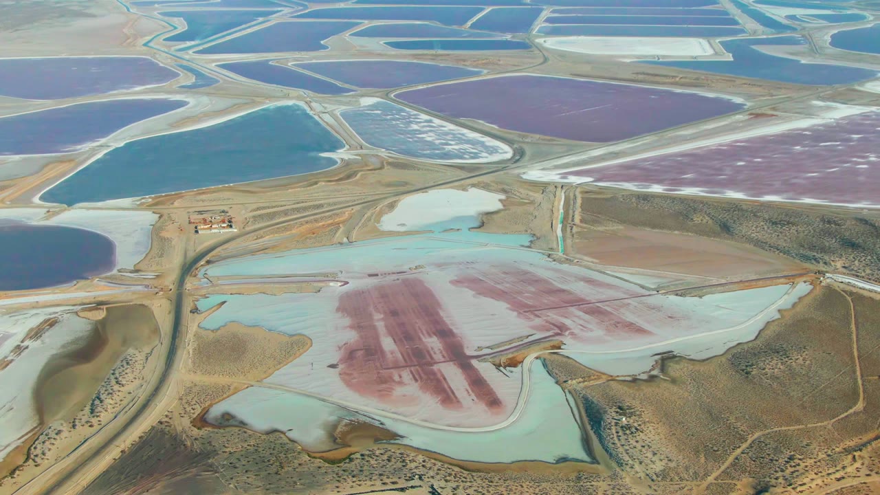 Aerial view over Guerrero Negro salt evaporation ponds with blue, purple, pink, and pale turquoise basins, dry roads, arid ground, and no visible people.