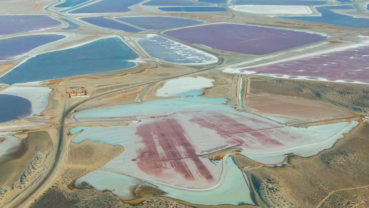 A drone-like aerial view over Guerrero Negro shows industrial salt ponds in blue, purple, pink, and pale green tones, with straight roads, dry terrain, and no visible people.