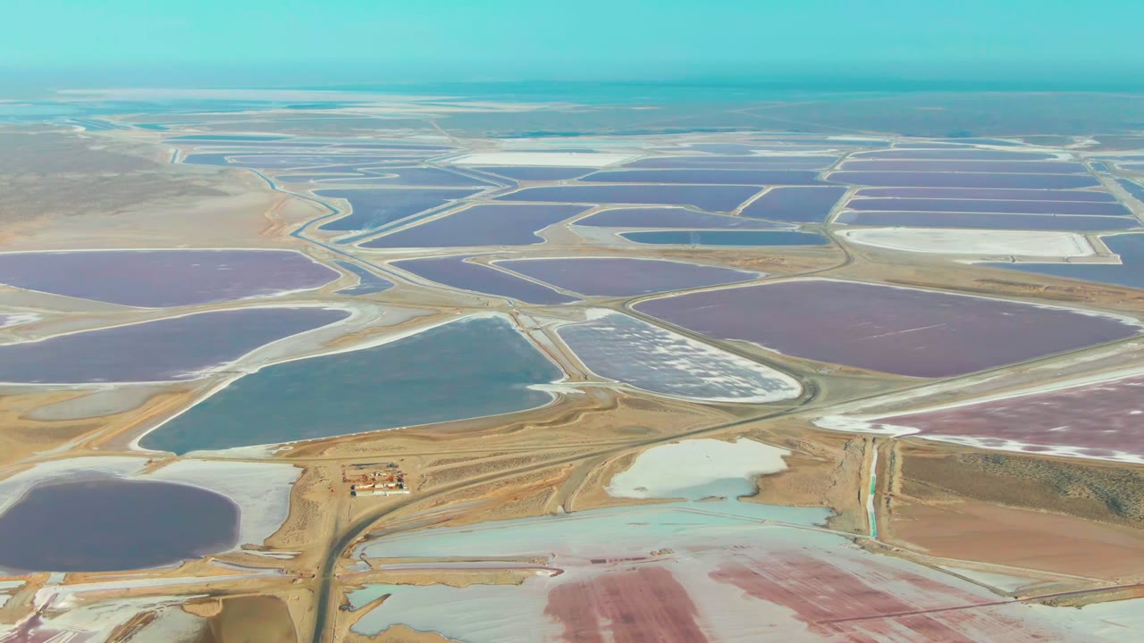 Oblique aerial view over Guerrero Negro salt evaporation ponds, with blue, purple, pink, white, and pale green basins divided by tan earth berms and no visible people.