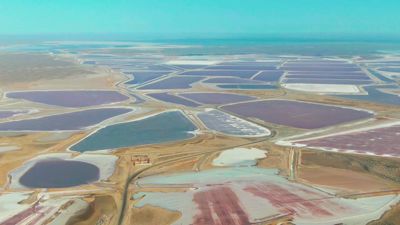 Guerrero Negro aerial view with a large teal pond in the lower center, purple and pink basins across the middle distance, white salt sections, and no visible people.