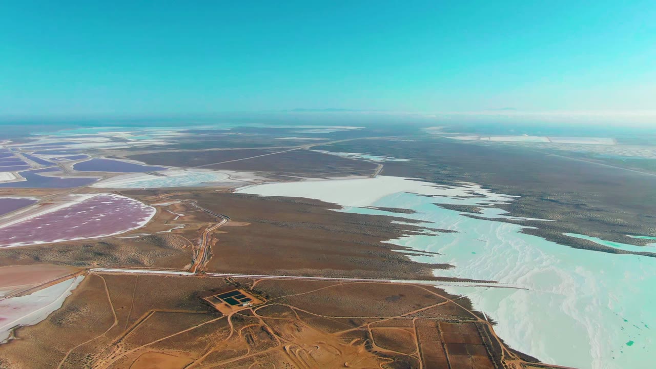Aerial view over Guerrero Negro salt evaporation ponds, with pale turquoise water at right, dry brown land and roads below, pink ponds in the distance, and no visible people.