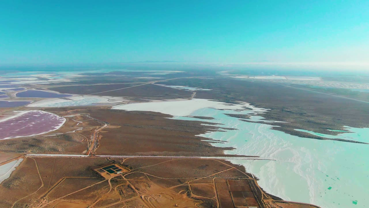 Opening aerial frame over Guerrero Negro salt lakes with turquoise water, white salt flats, brown desert roads, a pink basin at left, and no visible people.