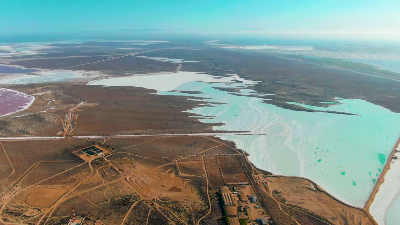 Opening aerial frame over Guerrero Negro, Baja California Sur, with turquoise and white salt ponds beside dry brown terrain, dirt roads, small structures, and no visible people.