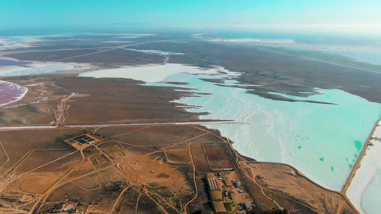 Mid-clip aerial view of Guerrero Negro salt ponds with turquoise water, white mineral flats, brown arid land, intersecting roads, and no visible people.