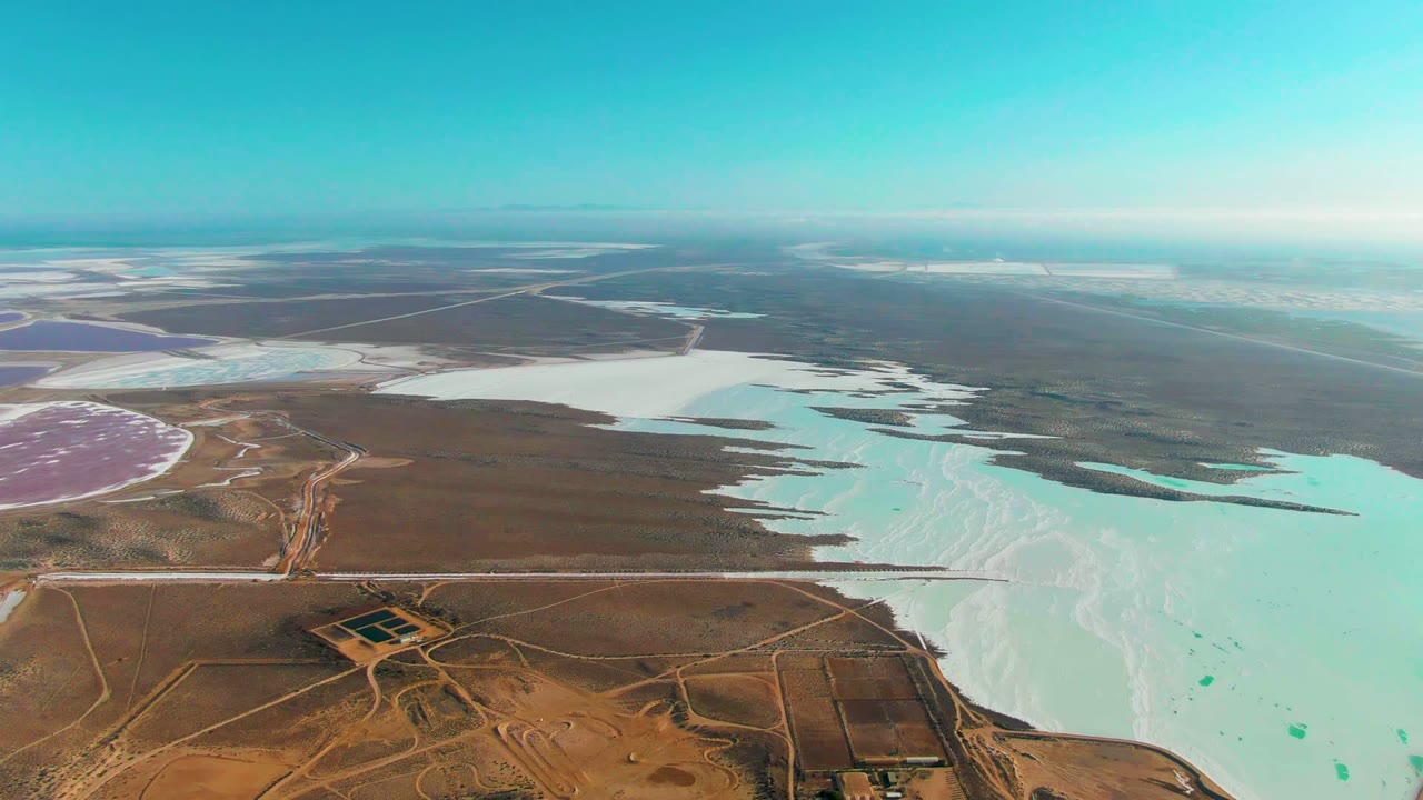 High aerial view over Guerrero Negro salt evaporation ponds, with pale blue-white water, brown desert roads, a pink basin at left, and no visible people.