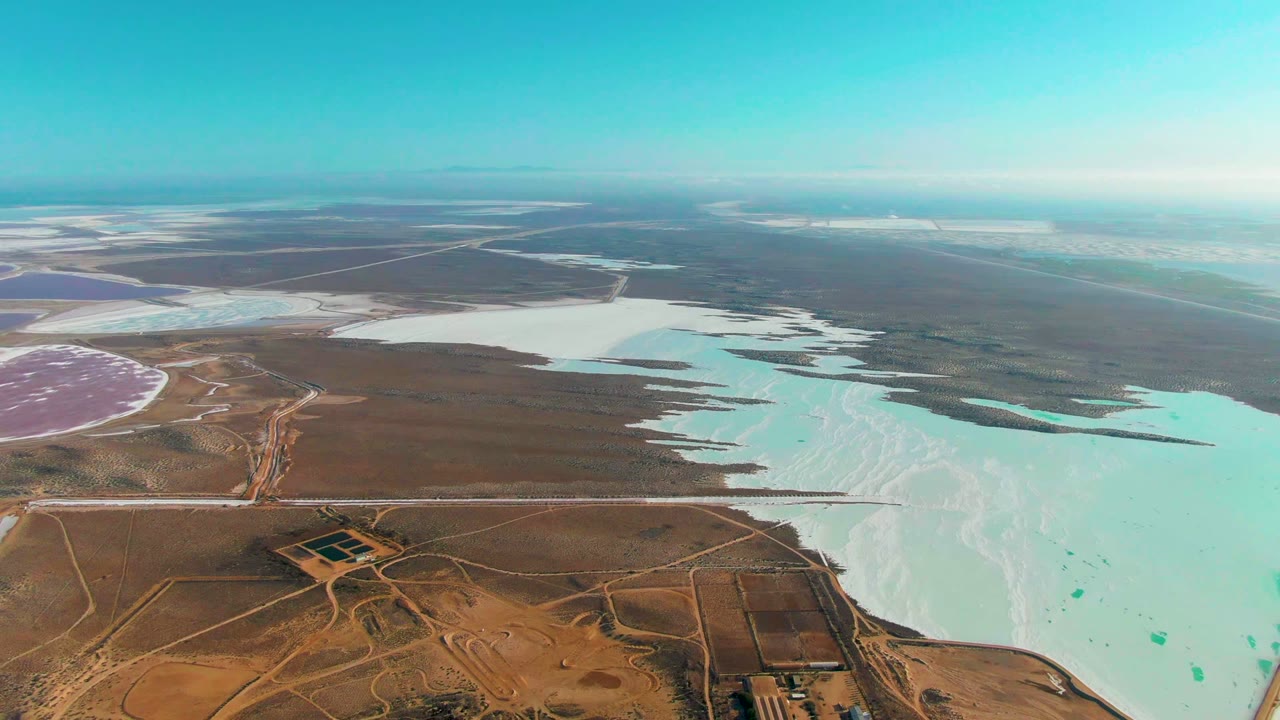 Aerial view across salt flats, turquoise pond edges, desert roads, and distant basins.