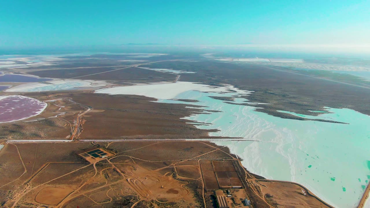 Wide aerial frame over Guerrero Negro with a large pale blue salt pond on the right, brown arid land below, a pink basin at left, distant white ponds, and no visible people.