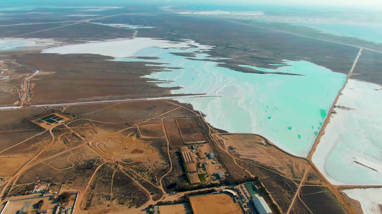 Opening aerial frame over Guerrero Negro salt flats with turquoise evaporation ponds, white salt deposits, dirt roads, industrial buildings, and no visible people.