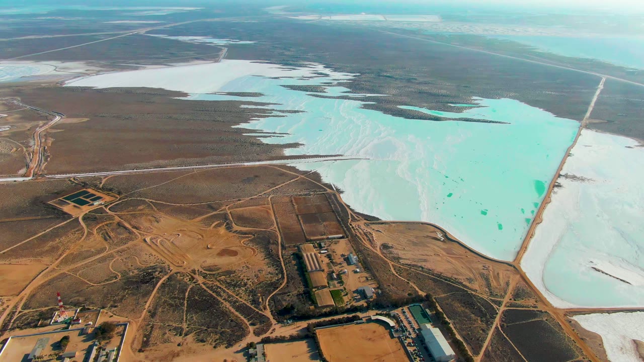 Aerial salt flat scene with turquoise ponds, white salt areas, roads, and small industrial structures.