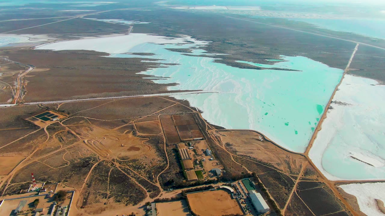 Third frame from the drone view over Guerrero Negro, with pale turquoise water filling the right midground, white salt deposits around it, industrial ponds at left, and no visible people.